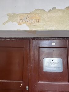 Górna część dużych, podwójnych drzwi z ciemnobrązowego drewna. Nad nimi widoczny fragment ściany, na którym spod odłupanej warstwy farby odsłonił się stary napis w języku polskim: „religia” – litery są lekko pochylone, w kolorze szaroczarnym na żółtawym tle. Farba wokół napisu jest w dużej części zdarta, pozostawiając nieregularne krawędzie. Na prawym skrzydle drzwi znajduje się prostokątna, metalowa tabliczka pamiątkowa w kolorze srebrnym z czarnym napisem: „SALA MATEMATYCZNA” wraz z informacjami o patronie i dacie. Tabliczka jest przykręcona czterema śrubami, po jednej w każdym rogu. Po lewej stronie na drzwiach widoczna jest niewielka kłódka lub element zamka.