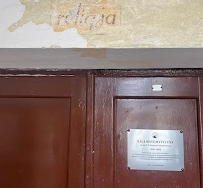 Górna część dużych, podwójnych drzwi z ciemnobrązowego drewna. Nad nimi widoczny fragment ściany, na którym spod odłupanej warstwy farby odsłonił się stary napis w języku polskim: „religia” – litery są lekko pochylone, w kolorze szaroczarnym na żółtawym tle. Farba wokół napisu jest w dużej części zdarta, pozostawiając nieregularne krawędzie. Na prawym skrzydle drzwi znajduje się prostokątna, metalowa tabliczka pamiątkowa w kolorze srebrnym z czarnym napisem: „SALA MATEMATYCZNA” wraz z informacjami o patronie i dacie. Tabliczka jest przykręcona czterema śrubami, po jednej w każdym rogu. Po lewej stronie na drzwiach widoczna jest niewielka kłódka lub element zamka.