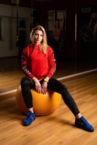 Zdjęcie przedstawia kobietę siedzącą na dużej, pomarańczowej piłce gimnastycznej w sali do ćwiczeń. Kobieta jest w centrum kadru, patrzy prosto w obiektyw z lekko poważnym, neutralnym wyrazem twarzy. Zdjęcie wykonane w profesjonalnym stylu z wyraźnym światłem skierowanym na postać – tło jest przyciemnione, a pierwszoplanowa postać silnie oświetlona, co uwydatnia szczegóły sylwetki i ubioru.

Szczegółowy opis postaci:

Pozycja ciała: Kobieta siedzi na pomarańczowej piłce fitness, stopy stabilnie ustawione na ziemi w lekkim rozkroku. Plecy proste, ramiona lekko opuszczone, dłonie swobodnie spoczywają na udach z palcami skierowanymi ku dołowi.

Wygląd zewnętrzny:

Cera jasna, twarz o delikatnych rysach, oczy jasne, podkreślone światłem.

Włosy długie, blond, rozpuszczone, lekko potargane i opadające na jedno ramię (prawe z perspektywy osoby na zdjęciu).

Paznokcie pomalowane na czerwono – dbałość o detale estetyczne.

Strój:

Bluza sportowa w intensywnym, czerwonym kolorze, z logiem marki „Hummel” na środku klatki piersiowej.

Rękawy bluzy zdobione charakterystycznym wzorem w kształcie białych strzałek (motyw marki Hummel), na czarnym tle – biegną wzdłuż całej długości rękawów.

Czarne legginsy sportowe, dopasowane do sylwetki, z lekkim połyskiem – w górnej części subtelny, prawie niewidoczny wzór.

Buty sportowe – intensywnie niebieskie sneakersy marki Nike, z czarnym logo „Swoosh” i czarną podeszwą.

Na lewym nadgarstku duży, żółty zegarek sportowy.

Tło i otoczenie:

Pomieszczenie:
Sala do ćwiczeń z drewnianą, jasnobrązową podłogą. Ściana po lewej stronie pokryta lustrem, co tworzy odbicie całej postaci – widoczne po prawej stronie kadru.

Tło:
Dość ciemne i mało oświetlone, ale widoczne są elementy siłowni:

Po lewej stronie – czarne sprzęty do ćwiczeń siłowych i taśmy TRX zwisające z sufitu.

W głębi, po prawej stronie – dwa duże plakaty:

Na jednym kobieta w sportowym staniku z rękami uniesionymi w górę (na plakacie napis „OMEGA” – prawdopodobnie reklama siłowni lub sprzętu).

Drugi plakat przedstawia zbliżenie na zaciśniętą pięść owiniętą w bandaż – sugeruje tematykę sportów walki.

W tle widoczne również niebieskie dozowniki z płynem (prawdopodobnie środki do dezynfekcji).

Kompozycja i styl zdjęcia:

Oświetlenie:
Silne światło padające z góry z lewej strony (z perspektywy widza), co tworzy kontrast między wyraźnie oświetloną postacią a przyciemnionym tłem.

Kąt zdjęcia:
Zdjęcie wykonane na wysokości oczu osoby siedzącej. Kadr symetryczny, centralnie skupiony na osobie.

Atmosfera:
Profesjonalna, sportowa, ale spokojna i statyczna. Zdjęcie emanuje poczuciem siły, kontroli i skupienia.