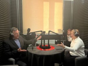 Zdjęcie przedstawia dwoje dorosłych – mężczyznę w garniturze i kobietę w białym swetrze – rozmawiających w profesjonalnym studiu radiowym. Oboje mają na uszach słuchawki i mówią do dużych mikrofonów na wysięgnikach. Siedzą przy okrągłym czarnym stole. Ściany wyłożone są dźwiękochłonną gąbką (czarne piankowe panele). Kobieta unosi ręce w ekspresyjnym geście, mężczyzna również gestykuluje żywo, oboje się uśmiechają. Na środku stołu znajduje się czerwony okrągły znak z napisem „STREFA KULTURY”.