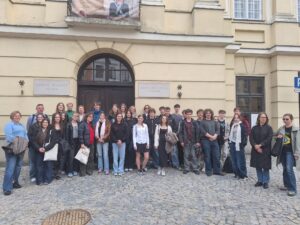 Zdjęcie przedstawia grupę około 30 osób (młodzież szkolna oraz kilku dorosłych) stojących na brukowanym placu przed zabytkowym, kremowym budynkiem z ciemnymi drewnianymi drzwiami. Nad wejściem widnieją dwie tablice z napisami: po lewej – "TRYBUNAŁ KORONNY 1578–1793", po prawej – "URZĄD STANU CYWILNEGO W LUBLINIE". Powyżej drzwi – baner z portretem duchownego. Wszyscy stoją równolegle do fasady, uśmiechają się. Część osób ubrana jest w kurtki, co może sugerować chłodniejszą porę roku. Kilka osób trzyma torby materiałowe.