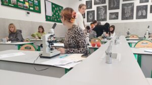 Przedstawia nowoczesną, jasną salę biologiczną. W pomieszczeniu stoi wiele długich, szarych stołów laboratoryjnych z zielonymi krzesłami. Na każdym stole znajduje się mikroskop oraz pojedyncze czerwone jabłko.
Na pierwszym planie siedzi dziewczyna w bluzce w cętki, z włosami upiętymi klamrą z pomarańczowym kwiatem. Patrzy w lewo, a przed nią leży mikroskop, kartka z rysunkiem komórek, długopis i telefon.
W tle widać inne uczennice i uczniów: niektórzy stoją, inni siedzą przy stołach, patrząc przez mikroskopy. Ściany ozdobione są czarno-białymi planszami anatomicznymi ukazującymi szkielety, organy i układ nerwowy. Z lewej strony ściany wiszą zielone tablice korkowe z kolorowymi zdjęciami i notatkami.