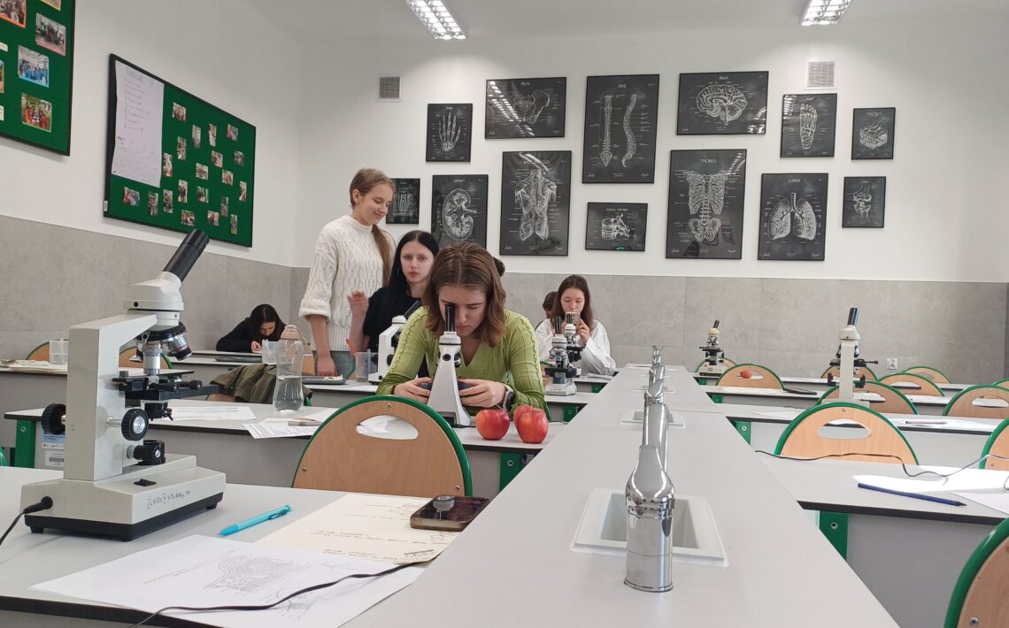 Ta sama sala ujęta z innej perspektywy. W centralnej części dziewczyna w jasnozielonym swetrze pochyla się nad mikroskopem, a obok niej na stole leżą dwa czerwone jabłka. Za nią stoją trzy uczennice rozmawiające; jedna z nich ma biały sweter, druga czarne włosy. W tle kolejne osoby pracują przy mikroskopach. Ściana naprzeciwko ozdobiona jest czarno-białymi planszami anatomicznymi z podpisami: „brain” (mózg), „lungs” (płuca), „thorax” (klatka piersiowa). Pomieszczenie jest dobrze oświetlone światłem jarzeniowym.