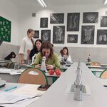 Ta sama sala ujęta z innej perspektywy. W centralnej części dziewczyna w jasnozielonym swetrze pochyla się nad mikroskopem, a obok niej na stole leżą dwa czerwone jabłka. Za nią stoją trzy uczennice rozmawiające; jedna z nich ma biały sweter, druga czarne włosy. W tle kolejne osoby pracują przy mikroskopach. Ściana naprzeciwko ozdobiona jest czarno-białymi planszami anatomicznymi z podpisami: „brain” (mózg), „lungs” (płuca), „thorax” (klatka piersiowa). Pomieszczenie jest dobrze oświetlone światłem jarzeniowym.