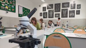 Kadr bliższy na mikroskop i uczniów. Na pierwszym planie po lewej stronie znajduje się mikroskop z widocznymi trzema obiektywami i pokrętłami regulacyjnymi. W tle widać tych samych uczniów, skupionych na obserwacji.
Dziewczyna w jasnozielonym swetrze spogląda w mikroskop, obok niej siedzi koleżanka z długim jasnym warkoczem. Po prawej stronie chłopak w białej koszulce pochyla się nad stołem, rozmawiając z kolegą.