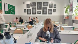 Widok sali od strony innego stołu. Na pierwszym planie uczennica w szarej, luźnej bluzie siedzi przy mikroskopie i patrzy przez jego okular.
W tle kilka grup uczniów pracuje – niektórzy siedzą, inni stoją, rozmawiają lub robią notatki. Na ścianie z tyłu wyraźnie widoczne są plansze z anatomicznymi rysunkami mózgu, kręgosłupa, układu nerwowego i szkieletu.
Po prawej stronie parapet z kilkoma dużymi doniczkami z zielonymi roślinami.