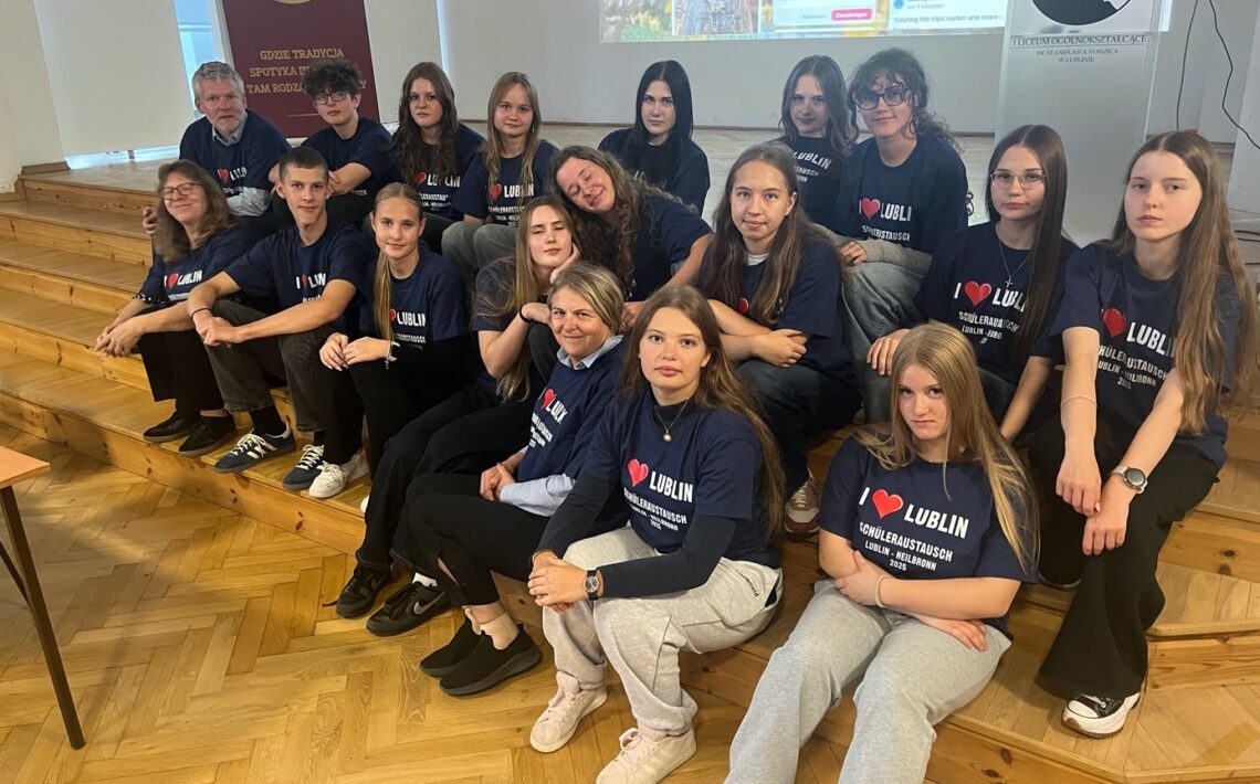 Zdjęcie grupowe młodzieży w sali szkolnej. Około siedemnaście osób – młodzież i dwoje nauczycieli – siedzi na drewnianych schodkach w sali o jasnych ścianach. Wszyscy mają granatowe koszulki z białym napisem: „I ♥ LUBLIN – SCHÜLERAUSTAUSCH LUBLIN – HEILBRONN 2024”. W tle widać rzutnik z prezentacją oraz szkolny baner z logo i napisem o tradycji szkoły. Osoby patrzą w stronę aparatu, niektóre się uśmiechają, panuje atmosfera wspólnoty i dumy z projektu wymiany.