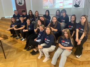 Zdjęcie grupowe młodzieży w sali szkolnej.
Około siedemnaście osób – młodzież i dwoje nauczycieli – siedzi na drewnianych schodkach w sali o jasnych ścianach.
Wszyscy mają granatowe koszulki z białym napisem:
„I ♥ LUBLIN – SCHÜLERAUSTAUSCH LUBLIN – HEILBRONN 2024”.
W tle widać rzutnik z prezentacją oraz szkolny baner z logo i napisem o tradycji szkoły.
Osoby patrzą w stronę aparatu, niektóre się uśmiechają, panuje atmosfera wspólnoty i dumy z projektu wymiany.