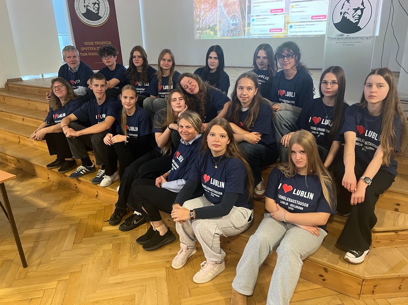Zdjęcie grupowe młodzieży w sali szkolnej. Około siedemnaście osób – młodzież i dwoje nauczycieli – siedzi na drewnianych schodkach w sali o jasnych ścianach. Wszyscy mają granatowe koszulki z białym napisem: „I ♥ LUBLIN – SCHÜLERAUSTAUSCH LUBLIN – HEILBRONN 2024”. W tle widać rzutnik z prezentacją oraz szkolny baner z logo i napisem o tradycji szkoły. Osoby patrzą w stronę aparatu, niektóre się uśmiechają, panuje atmosfera wspólnoty i dumy z projektu wymiany.
