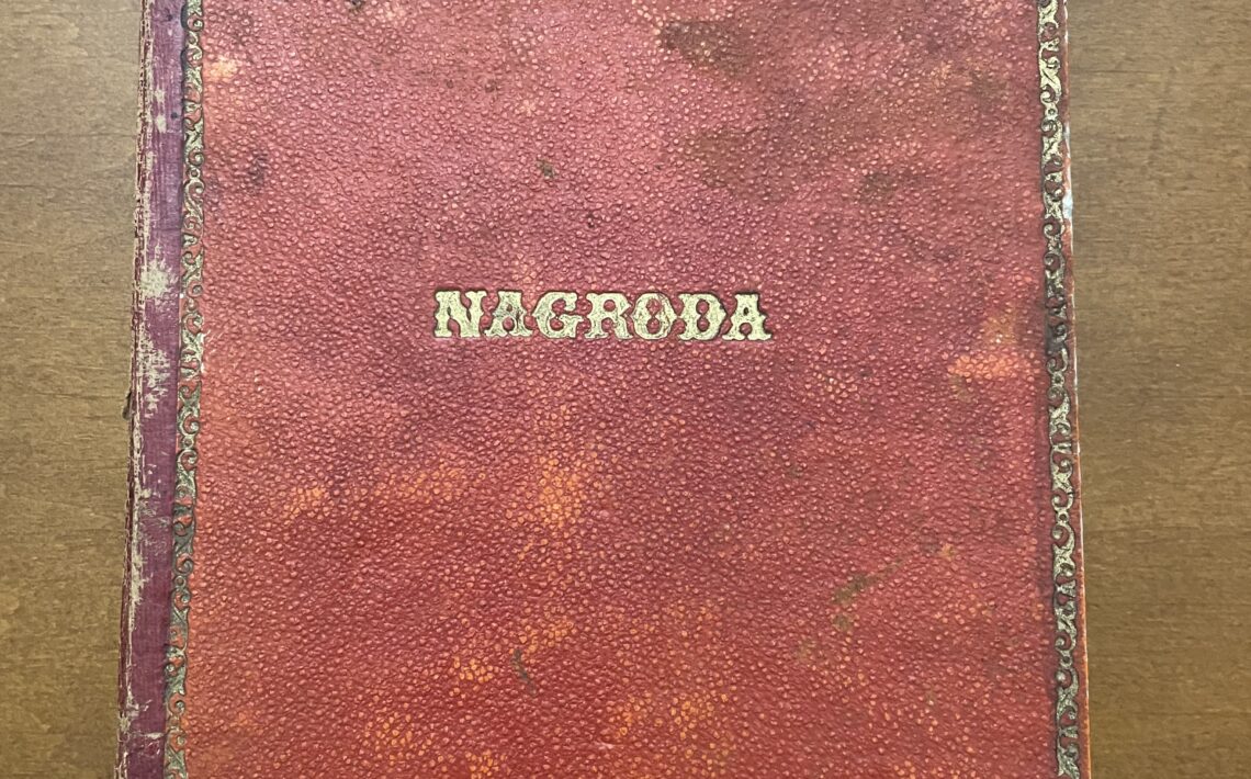 Pokazana jest okładka książki w kolorze ciemnoczerwonym, miejscami przetartym do koloru pomarańczowego. W centrum okładki, złotymi literami, znajduje się napis „NAGRODA” (drukowanymi literami). Krawędzie okładki zdobi cienka złota ramka z delikatnym ornamentem. Na górze i w dolnej części widać ślady zużycia – zagięcia, przetarcia, plamy i ubytki farby. Książka leży na brązowym, gładkim blacie.