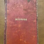 Pokazana jest okładka książki w kolorze ciemnoczerwonym, miejscami przetartym do koloru pomarańczowego. W centrum okładki, złotymi literami, znajduje się napis „NAGRODA” (drukowanymi literami). Krawędzie okładki zdobi cienka złota ramka z delikatnym ornamentem. Na górze i w dolnej części widać ślady zużycia – zagięcia, przetarcia, plamy i ubytki farby. Książka leży na brązowym, gładkim blacie.