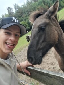 Zbliżenie przedstawiające młodą osobę robiącą selfie z koniem.
Po lewej stronie widoczna część twarzy uśmiechniętej osoby w czapce z daszkiem i beżowej bluzie z kapturem. Po prawej stronie – głowa ciemnobrązowego konia z rozwianą grzywą i wyraźnym okiem.
W tle zielona trawa i drzewa, prawdopodobnie wybieg dla koni. Zdjęcie ma przyjazny, spontaniczny charakter.