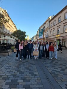 Zdjęcie grupowe na ulicy miasta, prawdopodobnie w Lublinie.
Około dwudziestoosobowa grupa młodzieży stoi na brukowanym deptaku między kamienicami. Budynki po bokach mają jasne elewacje i wystawy sklepowe. Na niebie bezchmurne błękitne niebo, słońce nisko, co wskazuje na późne popołudnie.
Młodzież pozuje do zdjęcia, większość uśmiechnięta, ubrana w bluzy, kurtki i jeansy. Atmosfera radosna i towarzyska.