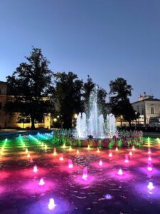 Wieczorne zdjęcie kolorowej fontanny na miejskim placu.
Na pierwszym planie widać płaską fontannę z kilkudziesięcioma strumieniami wody podświetlonymi intensywnymi kolorami: różowym, fioletowym, czerwonym, żółtym, zielonym i niebieskim. Woda odbija światła, tworząc efekt tęczy.
W centralnej części znajduje się wyższa fontanna z silnymi strumieniami wody unoszącymi się na około dwa metry, również podświetlonymi.
W tle – drzewa i oświetlone budynki miejskie. Nad całością ciemniejące niebo o barwie przechodzącej z błękitu w granat.