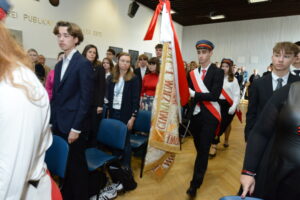Wnętrze dużej sali szkolnej o jasnych ścianach i drewnianej podłodze. Na zdjęciu widać grupę uczniów stojących w rzędach między niebieskimi krzesłami. Uczniowie są elegancko ubrani – chłopcy w ciemnych garniturach, dziewczęta w białych koszulach i ciemnych spódnicach lub spodniach.
Na pierwszym planie po prawej stronie idzie poczet sztandarowy – trzech uczniów: chłopiec niesie sztandar szkoły, a dwie dziewczęta w białych koszulach, czarnych spódnicach i czapkach z czerwonym otokiem towarzyszą mu z obu stron. Mają na sobie czerwono-białe szarfy i białe rękawiczki. Na sztandarze widnieją złote hafty i napis „GIMNAZJUM IM. STASZICA”. Wszyscy stoją poważni, skupieni, w tle widać nauczycieli i uczniów.
