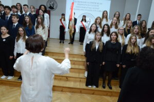 Grupa kilkudziesięciu uczniów stoi na schodkowej estradzie, śpiewając hymn lub pieśń szkolną. Wszyscy ubrani są odświętnie – dziewczęta w białych bluzkach i ciemnych spódnicach lub spodniach, chłopcy w garniturach i krawatach. Przed nimi stoi nauczycielka dyrygująca chórem – widoczna od tyłu, w białej bluzce.
Na tylnej ścianie widać duży ekran z napisem: „ŚLUBOWANIE UCZNIÓW KLAS PIERWSZYCH I LICEUM OGÓLNOKSZTAŁCĄCEGO IM. STANISŁAWA STASZICA W LUBLINIE” oraz okrągłe logo szkoły z profilem Stanisława Staszica. W tle stoi poczet sztandarowy z flagą i uczniowie w czerwonych szarfach.