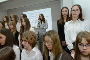 Zbliżenie na grupę uczennic stojących w pierwszym rzędzie chóru. Na środku za przezroczystą mównicą przemawia młoda uczennica w jasnym żakiecie w paski, trzyma mikrofon. Wokół niej stoją uczniowie w białych koszulach, niektórzy w czarnych marynarkach. W tle widoczny napis jak wcześniej oraz logo szkoły. Dziewczęta mają skupione, poważne twarze, niektóre trzymają ręce splecione przed sobą.