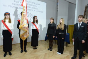 Na scenie stoi poczet sztandarowy – trójka uczniów w białych koszulach, czarnych długich spódnicach i granatowych czapkach z czerwonym paskiem, w czerwono-białych szarfach i białych rękawiczkach. W centrum chłopiec trzyma pionowo sztandar. Naprzeciw nich, w jednym rzędzie, stoją czterej uczniowie w ciemnych garniturach i eleganckich strojach, z poważnymi minami. Na ścianie za nimi ten sam napis i logo szkoły. Scena oświetlona jasnym światłem.