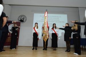 Moment ślubowania. Poczet sztandarowy stoi nieruchomo pośrodku sceny, a uczennice i uczniowie stojący naprzeciw wyciągają prawe ręce przed siebie, w geście przysięgi. Wszyscy są ubrani galowo. W tle biały ekran z napisem uroczystości oraz godło szkoły. Gest jest synchroniczny, symbolizujący powagę ceremonii.