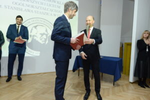 Na scenie dwóch mężczyzn w garniturach wymienia się dokumentami. Ten po prawej stronie, z krótkim zarostem i czerwoną krawatą, wręcza dyplom drugiemu, który trzyma go obiema rękami. W tle widoczny ekran z logo szkoły oraz osoby czekające na swoją kolej – stoją w rzędzie i trzymają teczki.