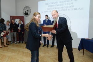 Mężczyzna w garniturze i czerwonej krawacie wręcza dokument kobiecie w ciemnej marynarce i okularach. Kobieta uśmiecha się lekko i podaje mu rękę. W tle widać grupę nauczycieli w eleganckich strojach, trzymających czerwone teczki. Wszyscy stoją w rządzie przy ścianie z logo szkoły.