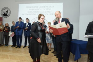 Mężczyzna w garniturze i czerwonej krawacie wręcza dyplom kobiecie w czarnej sukience z czerwonymi dodatkami – długim naszyjnikiem i bransoletkami. Kobieta ma krótkie włosy i czarne rajstopy, uśmiecha się lekko. W tle stoi grupa nauczycieli w różnym wieku, ubranych elegancko, z dyplomami w rękach. Widać serdeczną, uroczystą atmosferę.