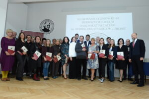 Zdjęcie grupowe. Przed sceną, na drewnianej podłodze, stoi duża grupa nauczycieli i pracowników szkoły – około 25 osób. Wszyscy trzymają w rękach czerwono-beżowe teczki z herbem szkoły. Z prawej strony stoi mężczyzna w ciemnym garniturze i czerwonej krawacie. Kobiety ubrane są elegancko, w sukienkach, garniturach lub żakietach. Niektórzy uśmiechają się, inni zachowują powagę. W tle ekran z napisem o ślubowaniu uczniów i duże logo szkoły z profilem Stanisława Staszica.