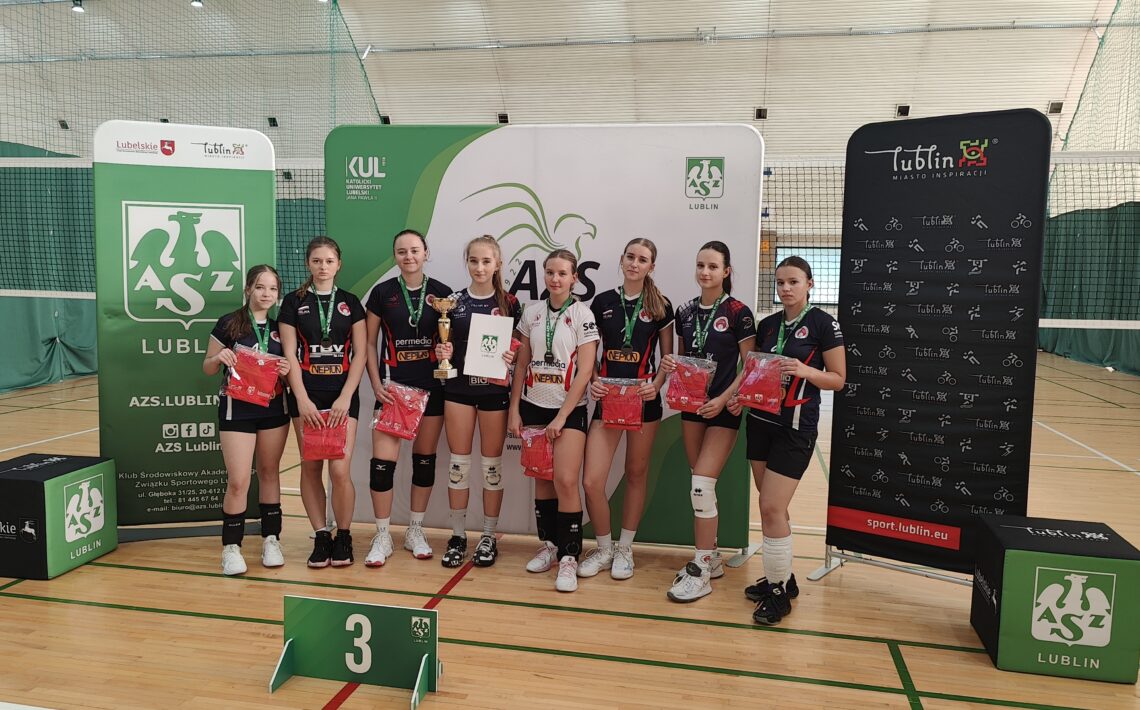 Na zdjęciu widać grupę ośmiu młodych siatkarek stojących w hali sportowej na parkiecie do siatkówki. Wszystkie mają na sobie stroje sportowe – czarne lub czarno-białe koszulki z krótkimi spodenkami, sportowe buty i białe lub czarne ochraniacze na kolana. Każda z zawodniczek ma na szyi zielony medal z białym logo. Zawodniczki stoją w szeregu i uśmiechają się do zdjęcia. W rękach trzymają czerwone torby lub koszulki – prawdopodobnie nagrody. Jedna z dziewcząt, stojąca mniej więcej w środku, trzyma złoty puchar w prawej ręce, a w lewej białą teczkę lub dyplom z logo organizatora. Za drużyną stoją trzy duże ścianki promocyjne w kolorach zieleni, bieli i czerni. Na środkowej i lewej widnieje duże zielone logo AZS Lublin – stylizowany biały gryf z rozpostartymi skrzydłami i literami „A Z S” pod spodem. Widoczne są również napisy: „AZS Lublin”, „KUL – Katolicki Uniwersytet Lubelski Jana Pawła II”, „Lubelskie”, oraz adresy internetowe i ikony mediów społecznościowych. Po prawej stronie ścianka jest czarna z białymi piktogramami różnych dyscyplin sportowych i czerwonym napisem „Lublin – Miasto Inspiracji” oraz adresem „sport.lublin.eu”. Przed zawodniczkami, na podłodze, znajduje się zielony stojak z białą cyfrą „3”, co oznacza, że drużyna zajęła trzecie miejsce w zawodach. Tło zdjęcia stanowi siatka do siatkówki i zielone osłony ścian hali. Oświetlenie jest jasne i równomierne – typowe dla sali gimnastycznej. Całość przedstawia moment dekoracji sportowej – młode siatkarki po zakończonym turnieju otrzymują nagrody i pozują do pamiątkowego zdjęcia.
