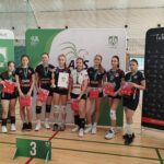 Na zdjęciu widać grupę ośmiu młodych siatkarek stojących w hali sportowej na parkiecie do siatkówki. Wszystkie mają na sobie stroje sportowe – czarne lub czarno-białe koszulki z krótkimi spodenkami, sportowe buty i białe lub czarne ochraniacze na kolana. Każda z zawodniczek ma na szyi zielony medal z białym logo. Zawodniczki stoją w szeregu i uśmiechają się do zdjęcia. W rękach trzymają czerwone torby lub koszulki – prawdopodobnie nagrody. Jedna z dziewcząt, stojąca mniej więcej w środku, trzyma złoty puchar w prawej ręce, a w lewej białą teczkę lub dyplom z logo organizatora. Za drużyną stoją trzy duże ścianki promocyjne w kolorach zieleni, bieli i czerni. Na środkowej i lewej widnieje duże zielone logo AZS Lublin – stylizowany biały gryf z rozpostartymi skrzydłami i literami „A Z S” pod spodem. Widoczne są również napisy: „AZS Lublin”, „KUL – Katolicki Uniwersytet Lubelski Jana Pawła II”, „Lubelskie”, oraz adresy internetowe i ikony mediów społecznościowych. Po prawej stronie ścianka jest czarna z białymi piktogramami różnych dyscyplin sportowych i czerwonym napisem „Lublin – Miasto Inspiracji” oraz adresem „sport.lublin.eu”. Przed zawodniczkami, na podłodze, znajduje się zielony stojak z białą cyfrą „3”, co oznacza, że drużyna zajęła trzecie miejsce w zawodach. Tło zdjęcia stanowi siatka do siatkówki i zielone osłony ścian hali. Oświetlenie jest jasne i równomierne – typowe dla sali gimnastycznej. Całość przedstawia moment dekoracji sportowej – młode siatkarki po zakończonym turnieju otrzymują nagrody i pozują do pamiątkowego zdjęcia.