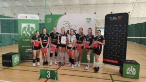 Na zdjęciu widać grupę ośmiu młodych siatkarek stojących w hali sportowej na parkiecie do siatkówki. Wszystkie mają na sobie stroje sportowe – czarne lub czarno-białe koszulki z krótkimi spodenkami, sportowe buty i białe lub czarne ochraniacze na kolana. Każda z zawodniczek ma na szyi zielony medal z białym logo.

Zawodniczki stoją w szeregu i uśmiechają się do zdjęcia. W rękach trzymają czerwone torby lub koszulki – prawdopodobnie nagrody. Jedna z dziewcząt, stojąca mniej więcej w środku, trzyma złoty puchar w prawej ręce, a w lewej białą teczkę lub dyplom z logo organizatora.

Za drużyną stoją trzy duże ścianki promocyjne w kolorach zieleni, bieli i czerni. Na środkowej i lewej widnieje duże zielone logo AZS Lublin – stylizowany biały gryf z rozpostartymi skrzydłami i literami „A Z S” pod spodem. Widoczne są również napisy:

„AZS Lublin”,

„KUL – Katolicki Uniwersytet Lubelski Jana Pawła II”,

„Lubelskie”,

oraz adresy internetowe i ikony mediów społecznościowych.

Po prawej stronie ścianka jest czarna z białymi piktogramami różnych dyscyplin sportowych i czerwonym napisem „Lublin – Miasto Inspiracji” oraz adresem „sport.lublin.eu”.

Przed zawodniczkami, na podłodze, znajduje się zielony stojak z białą cyfrą „3”, co oznacza, że drużyna zajęła trzecie miejsce w zawodach.

Tło zdjęcia stanowi siatka do siatkówki i zielone osłony ścian hali. Oświetlenie jest jasne i równomierne – typowe dla sali gimnastycznej.

Całość przedstawia moment dekoracji sportowej – młode siatkarki po zakończonym turnieju otrzymują nagrody i pozują do pamiątkowego zdjęcia.