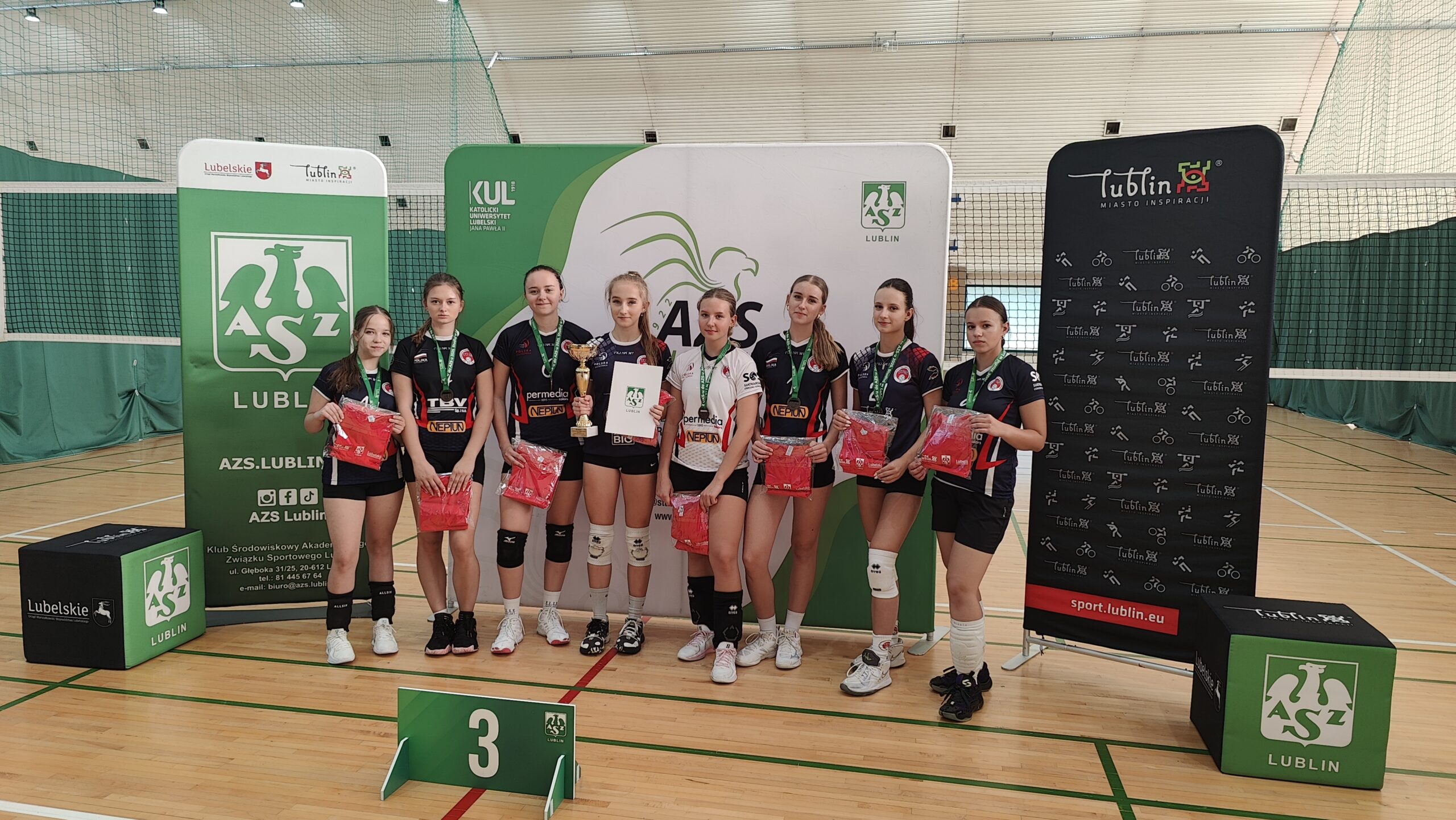 Na zdjęciu widać grupę ośmiu młodych siatkarek stojących w hali sportowej na parkiecie do siatkówki. Wszystkie mają na sobie stroje sportowe – czarne lub czarno-białe koszulki z krótkimi spodenkami, sportowe buty i białe lub czarne ochraniacze na kolana. Każda z zawodniczek ma na szyi zielony medal z białym logo. Zawodniczki stoją w szeregu i uśmiechają się do zdjęcia. W rękach trzymają czerwone torby lub koszulki – prawdopodobnie nagrody. Jedna z dziewcząt, stojąca mniej więcej w środku, trzyma złoty puchar w prawej ręce, a w lewej białą teczkę lub dyplom z logo organizatora. Za drużyną stoją trzy duże ścianki promocyjne w kolorach zieleni, bieli i czerni. Na środkowej i lewej widnieje duże zielone logo AZS Lublin – stylizowany biały gryf z rozpostartymi skrzydłami i literami „A Z S” pod spodem. Widoczne są również napisy: „AZS Lublin”, „KUL – Katolicki Uniwersytet Lubelski Jana Pawła II”, „Lubelskie”, oraz adresy internetowe i ikony mediów społecznościowych. Po prawej stronie ścianka jest czarna z białymi piktogramami różnych dyscyplin sportowych i czerwonym napisem „Lublin – Miasto Inspiracji” oraz adresem „sport.lublin.eu”. Przed zawodniczkami, na podłodze, znajduje się zielony stojak z białą cyfrą „3”, co oznacza, że drużyna zajęła trzecie miejsce w zawodach. Tło zdjęcia stanowi siatka do siatkówki i zielone osłony ścian hali. Oświetlenie jest jasne i równomierne – typowe dla sali gimnastycznej. Całość przedstawia moment dekoracji sportowej – młode siatkarki po zakończonym turnieju otrzymują nagrody i pozują do pamiątkowego zdjęcia.