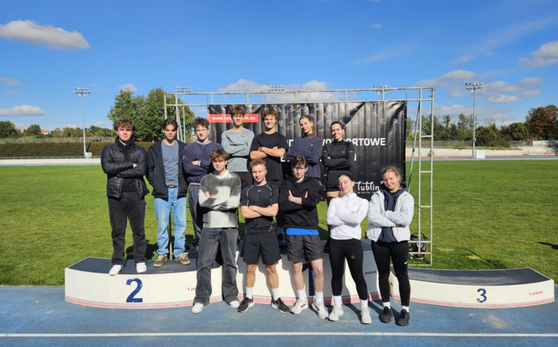 Na zdjęciu znajduje się grupa młodych osób – prawdopodobnie uczniów lub studentów – stojących na podium sportowym na stadionie lekkoatletycznym. Zdjęcie wykonano w pogodny, słoneczny dzień, pod błękitnym niebem z kilkoma białymi chmurkami. Opis szczegółowy: W centrum obrazu znajduje się podium z trzema stopniami oznaczonymi cyframi 2 (po lewej), 1 (środkowy, niewidoczny – zasłonięty przez grupę) i 3 (po prawej). Grupa składa się z 12 osób – 8 mężczyzn i 4 kobiet. Pierwszy rząd (na dole, stojący na podium): Pięć osób stoi z rękami skrzyżowanymi na piersiach. Od lewej: młody mężczyzna w czarnej kurtce i ciemnych spodniach stoi na miejscu oznaczonym cyfrą 2; obok niego dwóch chłopaków w sportowych ubraniach; po prawej stronie trzy młode kobiety w sportowych strojach – legginsy, bluzy, jedna z nich w białej kurtce. Wszyscy mają na sobie sportowe buty. Drugi rząd (z tyłu, na ziemi lub wyższym poziomie podium): Siedem osób – głównie chłopcy – stoi w lekkim rozluźnieniu, część z rękami skrzyżowanymi, część trzyma je swobodnie. Wszyscy patrzą w stronę aparatu i uśmiechają się lekko lub zachowują neutralny wyraz twarzy. Ubrani są w sportowe bluzy, dresy, kurtki lub swetry. Za grupą widoczny jest czarny baner z białym napisem: „www.sport.lublin.eu”