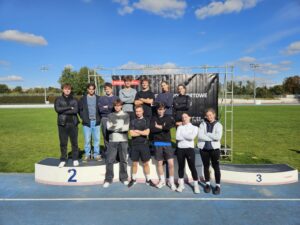 Na zdjęciu znajduje się grupa młodych osób – prawdopodobnie uczniów lub studentów – stojących na podium sportowym na stadionie lekkoatletycznym. Zdjęcie wykonano w pogodny, słoneczny dzień, pod błękitnym niebem z kilkoma białymi chmurkami.

Opis szczegółowy:
W centrum obrazu znajduje się podium z trzema stopniami oznaczonymi cyframi 2 (po lewej), 1 (środkowy, niewidoczny – zasłonięty przez grupę) i 3 (po prawej). Grupa składa się z 12 osób – 8 mężczyzn i 4 kobiet.

Pierwszy rząd (na dole, stojący na podium):
Pięć osób stoi z rękami skrzyżowanymi na piersiach. Od lewej: młody mężczyzna w czarnej kurtce i ciemnych spodniach stoi na miejscu oznaczonym cyfrą 2; obok niego dwóch chłopaków w sportowych ubraniach; po prawej stronie trzy młode kobiety w sportowych strojach – legginsy, bluzy, jedna z nich w białej kurtce. Wszyscy mają na sobie sportowe buty.

Drugi rząd (z tyłu, na ziemi lub wyższym poziomie podium):
Siedem osób – głównie chłopcy – stoi w lekkim rozluźnieniu, część z rękami skrzyżowanymi, część trzyma je swobodnie. Wszyscy patrzą w stronę aparatu i uśmiechają się lekko lub zachowują neutralny wyraz twarzy. Ubrani są w sportowe bluzy, dresy, kurtki lub swetry.

Za grupą widoczny jest czarny baner z białym napisem:
„www.sport.lublin.eu”
