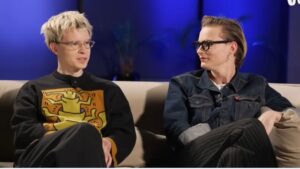 Na zdjęciu widać dwóch młodych mężczyzn siedzących obok siebie na beżowej kanapie w dobrze oświetlonym pomieszczeniu. Tło jest ciemne, podświetlone intensywnym niebieskim światłem, które tworzy kontrast z jasnym pierwszym planem. W tle można dostrzec rozmazany zarys rośliny doniczkowej. Opis postaci po lewej stronie: Mężczyzna siedzi z nogą założoną na nogę. Ma krótkie, jasne włosy w odcieniu blond, lekko potargane. Nosi przezroczyste okulary w cienkich oprawkach. Ubrany jest w czarny sweter z dużym, kolorowym nadrukiem przedstawiającym stylizowaną, żółtą postać o grubych konturach, przypominającą grafikę w stylu Keitha Haringa. Postać ma uniesione ręce, a tło nadruku jest pomarańczowe z fioletowymi elementami. Mężczyzna ma założony mikrofon przypięty do swetra, co sugeruje, że uczestniczy w nagraniu wywiadu lub rozmowy. Opis postaci po prawej stronie: Mężczyzna siedzi obok, lekko zwrócony w stronę rozmówcy po lewej, z którym wymienia spojrzenie. Ma włosy w kolorze jasnobrązowym, zaczesane gładko do tyłu. Nosi ciemne, kwadratowe okulary w czarnych oprawkach oraz kolczyk w lewym uchu. Ubrany jest w jeansową kurtkę z widocznymi szwami i metalowymi guzikami, pod którą ma jasny T-shirt. Spodnie są ciemne, w drobne pionowe paski. Obaj wyglądają na skupionych i zaangażowanych w rozmowę. Atmosfera jest spokojna i profesjonalna, a ustawienie kanapy i oświetlenia wskazuje, że scena pochodzi z wywiadu lub programu telewizyjnego.