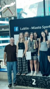 Zdjęcie przedstawia grupę młodych dziewcząt stojących na podium pływalni. W tle widoczna jest duża czarna tablica z napisem „Lublin – Miasto Sportu” oraz charakterystycznym znakiem Facebooka w kolorze niebieskim. Za tablicą znajdują się duże, nowoczesne przeszklenia basenu, przez które wpada naturalne światło.
Na podium stoją trzy dziewczęta, ubrane w luźne, sportowe ubrania.
Dziewczyna stojąca pośrodku (na miejscu drugim – oznaczonym białą cyfrą 2 na czarnym tle) ma na sobie czarno-szary strój sportowy z geometrycznym wzorem. W ręku trzyma złoty puchar, a na szyi ma srebrny medal. W drugiej dłoni trzyma biały dyplom.
Obok niej, po lewej stronie (patrząc od widza), stoi dziewczyna w czarnym topie i szerokich, szarych spodniach w kratę. Na szyi ma medal i również trzyma biały dyplom.
Po prawej stronie podium stoi dziewczyna w szarej koszulce i ciemnych szerokich spodniach, z medalem na szyi i lekko pochyloną głową w uśmiechu.
Za nimi widać kilka innych dziewcząt, również trzymających dyplomy, częściowo zasłoniętych przez stojące z przodu zawodniczki.
Po lewej stronie zdjęcia stoi dorosły mężczyzna – prawdopodobnie trener lub organizator zawodów. Ubrany jest w brązową koszulkę polo i niebieskie spodnie. Na nogach ma czarne klapki. Uśmiecha się lekko i patrzy w stronę aparatu.
Na górze po lewej stronie kadru widoczny jest duży zegar sportowy z czerwonymi, żółtymi i czarnymi wskazówkami.
Scena przedstawia moment wręczenia nagród lub zakończenia zawodów pływackich – uczestniczki pozują do pamiątkowego zdjęcia na podium.