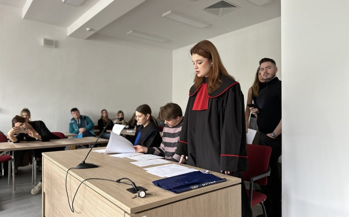 Na zdjęciu znajduje się scena rozgrywająca się w pomieszczeniu przypominającym salę sądową lub salę symulacji rozprawy — prawdopodobnie na uczelni. Na pierwszym planie widać dużą, jasnobrązową, drewnianą ławę (biurko) ustawioną poziomo względem widza. Na biurku leży kilka arkuszy białych dokumentów, teczka w granatowym kolorze oraz mikrofon na elastycznym przewodzie, skierowany w stronę osoby stojącej. Przy biurku siedzą dwie osoby. Kobieta po lewej stronie ma gładko zaczesane, ciemne włosy spięte z tyłu głowy, ubrana jest w czarny strój z niebieskim żabotem — przypominający togę sędziowską lub adwokacką. Przegląda dokumenty, lekko pochylona do przodu. Obok niej, po prawej stronie, siedzi mężczyzna w różowo-szarej, poziomej, prążkowanej bluzie. On również ma przed sobą dokumenty, które czyta z lekkim pochyleniem głowy. Po prawej stronie, na stojąco, znajduje się młoda kobieta w czarnej todze z czerwonymi lamówkami i dużym czerwonym żabotem. Ma długie, rude włosy rozpuszczone, a jej twarz jest poważna i skupiona. Wydaje się, że przemawia lub odczytuje coś z kartki znajdującej się na biurku. W tle, przy ścianie po lewej stronie, siedzi kilka osób — około sześć. Niektórzy obserwują sytuację z zainteresowaniem, inni rozmawiają między sobą lub przeglądają coś w rękach. Jedna kobieta z lewej strony wspiera głowę na dłoni, wygląda na znudzoną lub zmęczoną. Z prawej strony, w pobliżu ściany, stoją jeszcze dwie osoby — mężczyzna w czarnym golfie trzymający telefon w dłoni oraz kobieta w jasnobrązowej marynarce lub swetrze. Pomieszczenie ma białe ściany, sufit z prostokątnymi panelami świetlnymi LED, a podłoga jest szara. Krzesła są czerwone z czarnymi metalowymi nogami. Oświetlenie jest naturalne — z lewej strony, poza kadrem, prawdopodobnie znajduje się okno, które doświetla całą scenę. Całość wygląda jak ćwiczenia lub symulacja rozprawy sądowej z udziałem studentów prawa, prowadzona w sali akademickiej.