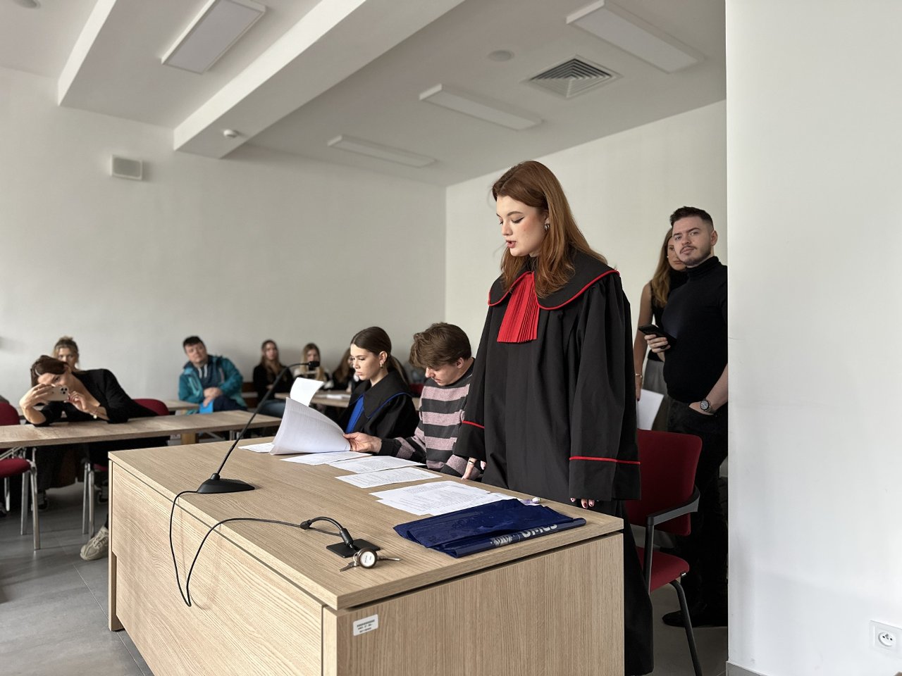 Na zdjęciu znajduje się scena rozgrywająca się w pomieszczeniu przypominającym salę sądową lub salę symulacji rozprawy — prawdopodobnie na uczelni. Na pierwszym planie widać dużą, jasnobrązową, drewnianą ławę (biurko) ustawioną poziomo względem widza. Na biurku leży kilka arkuszy białych dokumentów, teczka w granatowym kolorze oraz mikrofon na elastycznym przewodzie, skierowany w stronę osoby stojącej. Przy biurku siedzą dwie osoby. Kobieta po lewej stronie ma gładko zaczesane, ciemne włosy spięte z tyłu głowy, ubrana jest w czarny strój z niebieskim żabotem — przypominający togę sędziowską lub adwokacką. Przegląda dokumenty, lekko pochylona do przodu. Obok niej, po prawej stronie, siedzi mężczyzna w różowo-szarej, poziomej, prążkowanej bluzie. On również ma przed sobą dokumenty, które czyta z lekkim pochyleniem głowy. Po prawej stronie, na stojąco, znajduje się młoda kobieta w czarnej todze z czerwonymi lamówkami i dużym czerwonym żabotem. Ma długie, rude włosy rozpuszczone, a jej twarz jest poważna i skupiona. Wydaje się, że przemawia lub odczytuje coś z kartki znajdującej się na biurku. W tle, przy ścianie po lewej stronie, siedzi kilka osób — około sześć. Niektórzy obserwują sytuację z zainteresowaniem, inni rozmawiają między sobą lub przeglądają coś w rękach. Jedna kobieta z lewej strony wspiera głowę na dłoni, wygląda na znudzoną lub zmęczoną. Z prawej strony, w pobliżu ściany, stoją jeszcze dwie osoby — mężczyzna w czarnym golfie trzymający telefon w dłoni oraz kobieta w jasnobrązowej marynarce lub swetrze. Pomieszczenie ma białe ściany, sufit z prostokątnymi panelami świetlnymi LED, a podłoga jest szara. Krzesła są czerwone z czarnymi metalowymi nogami. Oświetlenie jest naturalne — z lewej strony, poza kadrem, prawdopodobnie znajduje się okno, które doświetla całą scenę. Całość wygląda jak ćwiczenia lub symulacja rozprawy sądowej z udziałem studentów prawa, prowadzona w sali akademickiej.