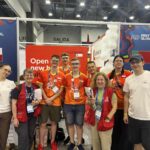 Grupowe zdjęcie kilku osób wykonane przy stoisku oznaczonym flagą Polski i napisem POLAND / POLONIA. W centrum stoi grupa młodych uczestników ubranych w pomarańczowo-czerwone koszulki z logo FIRST GLOBAL i biało-czerwonymi flagami na rękawach. Towarzyszy im kilka osób dorosłych – opiekunowie, mentorzy i organizatorzy. Dwie kobiety w czerwonych kamizelkach stoją z przodu i uśmiechają się. Po lewej stronie widoczny jest fragment innego stoiska, po prawej część stoiska z napisem Belarus. Tło stanowi ścianka wystawowa ozdobiona balonami i czerwonym banerem z hasłem „Open new horizons”. Widać też niebieskie identyfikatory na smyczach, które każdy z uczestników ma zawieszone na szyi. Zdjęcie przedstawia moment spotkania i wspólnej radości zespołu reprezentującego Polskę podczas międzynarodowych zawodów robotycznych FIRST GLOBAL, odbywających się w dużej hali targowej pełnej stoisk i uczestników z całego świata.