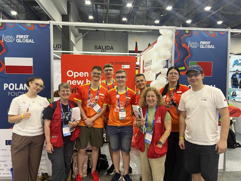 Grupowe zdjęcie kilku osób wykonane przy stoisku oznaczonym flagą Polski i napisem POLAND / POLONIA. W centrum stoi grupa młodych uczestników ubranych w pomarańczowo-czerwone koszulki z logo FIRST GLOBAL i biało-czerwonymi flagami na rękawach. Towarzyszy im kilka osób dorosłych – opiekunowie, mentorzy i organizatorzy. Dwie kobiety w czerwonych kamizelkach stoją z przodu i uśmiechają się. Po lewej stronie widoczny jest fragment innego stoiska, po prawej część stoiska z napisem Belarus. Tło stanowi ścianka wystawowa ozdobiona balonami i czerwonym banerem z hasłem „Open new horizons”. Widać też niebieskie identyfikatory na smyczach, które każdy z uczestników ma zawieszone na szyi. Zdjęcie przedstawia moment spotkania i wspólnej radości zespołu reprezentującego Polskę podczas międzynarodowych zawodów robotycznych FIRST GLOBAL, odbywających się w dużej hali targowej pełnej stoisk i uczestników z całego świata.