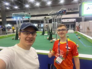 Na drugim zdjęciu widoczne są dwie osoby stojące przed areną zawodów robotycznych, uśmiechnięte i pozujące do wspólnego zdjęcia typu selfie. Osoba po lewej stronie ma czapkę z daszkiem, przezroczyste okulary ochronne i białą koszulkę. Po prawej stronie stoi uczestnik w pomarańczowo-czerwonej koszulce z logo FIRST GLOBAL oraz flagą Polski na rękawie. Ma zawieszony na szyi identyfikator i pokazuje uniesiony kciuk w geście zadowolenia. Za nimi znajduje się to samo zielone pole zawodów z konstrukcjami i kolorowymi piłkami. W tle widać transparent z napisem „EXIT” oraz inne stoiska uczestników. Zdjęcie oddaje radosny nastrój i poczucie dumy z uczestnictwa w międzynarodowym wydarzeniu naukowo-technicznym.