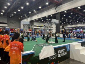 Zdjęcie przedstawia dużą halę wystawową o wysokim suficie i jasnym oświetleniu. Na środku znajduje się zielone pole z wyraźnie wyznaczonymi liniami i strukturami – to arena zawodów robotycznych. Na niej stoją wysokie, przezroczyste konstrukcje z metalu i plastiku, przypominające wieże lub bramki. Wokół nich porozrzucane są kolorowe piłki – głównie pomarańczowe, niebieskie i żółte. Na pierwszym planie, po lewej stronie, widać kilku młodych uczestników w pomarańczowo-czerwonych koszulkach z napisem na plecach „FIRST GLOBAL” i nazwą kraju Brunei Darussalam. Po prawej stronie areny stoi sędzia lub mentor ubrany w ciemną koszulkę i kamizelkę, obserwujący pole gry. W tle za areną widoczni są widzowie i stoiska wypełnione kolorowymi banerami i flagami. Nad wyjściem z hali widać duży napis „SALIDA / EXIT / EMERGENCIA”. Atmosfera zdjęcia sugeruje emocje i skupienie uczestników podczas międzynarodowych zawodów robotycznych.