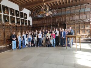 Fotografia przedstawia dużą grupę młodzieży i dorosłych – około trzydziestu osób – stojących w zabytkowym wnętrzu. Pomieszczenie jest bogato zdobione ciemnym, rzeźbionym drewnem. Na ścianach wisi rząd portretów przedstawiających mężczyzn w historycznych strojach, każdy w ozdobnej ramie. Sufit z drewnianych belek, a w centralnej części kadru zwisa ogromny żyrandol z metalowych elementów i herbów. Grupa stoi w półkolu, w centrum pomieszczenia, patrząc w stronę fotografa. Widać elementy wyposażenia: pulpit z drewna, balustrady i ozdobne panele. Zdjęcie ma charakter dokumentacyjny, prawdopodobnie wykonane podczas wizyty w muzeum lub zabytkowym ratuszu.