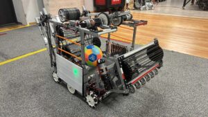 Na zdjęciu znajduje się złożony, zaawansowany robot mobilny, przypominający konstrukcję inżynierską używaną w zawodach robotycznych, np. typu FIRST Robotics. Opis szczegółowy, zgodny z zasadami dostępności WCAG 2.2: Robot ma kształt prostopadłościanu o wymiarach zbliżonych do średniego pudełka – mniej więcej 70 centymetrów długości, 50 centymetrów szerokości i 60 centymetrów wysokości. Konstrukcja wykonana jest głównie z aluminiowych profili w kształcie ceowników połączonych w sztywną ramę. Wewnątrz widoczne są liczne przewody, przekładnie, paski i silniki. Dolna część robota: Posiada cztery koła z przodu i z tyłu, wykonane z jasnoszarego tworzywa z licznymi rolkami bocznymi — to tzw. koła typu mecanum, umożliwiające ruch w dowolnym kierunku. Podłoże, na którym stoi robot, to wykładzina z krótkim włosiem w kolorze ciemnoszarym. Na jej krawędzi widać żółtą linię oddzielającą część pomieszczenia z podłogą drewnianą. Przód robota: Znajduje się tam mechanizm chwytający lub podający, złożony z kilku poziomych pasków gumowych naciągniętych na czarne koła zębate. Mechanizm jest lekko nachylony do przodu i przypomina ruchomy transporter taśmowy. Powyżej tej sekcji widać dwa pionowe pręty z czarnymi paskami gumowymi, prawdopodobnie do unoszenia piłki lub innego obiektu. Środkowa część robota: Wewnątrz konstrukcji znajduje się piłka o średnicy około 20 centymetrów, wielobarwna — składa się z sześciokątów w kolorach: pomarańczowym, żółtym, niebieskim i zielonym. Na piłce widoczny jest nadruk z napisem „GILBERT” i symbolem sportowym. Obok piłki można zauważyć liczne pomarańczowe pręty lub linki, które prawdopodobnie stabilizują mechanizmy wewnętrzne. Górna część robota: Umieszczono tu dwa duże, czarne silniki elektryczne z oznaczeniami „REV”, połączone z kołami napędowymi i rolkami. Prawdopodobnie służą do wyrzucania piłki z dużą prędkością. Całość ma bardzo techniczny wygląd – widać metalowe wsporniki, śruby i przewody prowadzone wzdłuż ramy. Dodatkowe szczegóły: Na jednym z boków świeci się zielona dioda LED, sygnalizująca prawdopodobnie zasilanie lub gotowość systemu. Tło zdjęcia przedstawia warsztat — drewnianą podłogę, metalowe wózki i czerwone narzędzia.