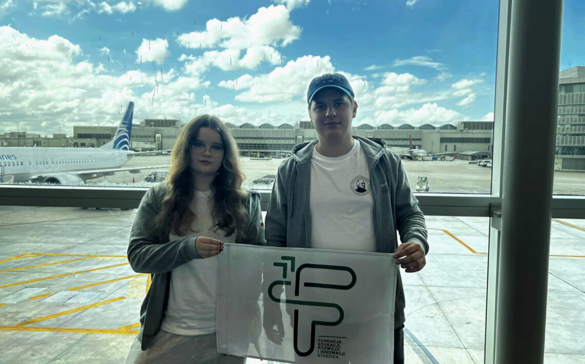 Opis szczegółowy zgodny z wymaganiami dostępności WCAG 2.2: Scena rozgrywa się w dzień, przy naturalnym świetle. Za dużą szybą w tle widać samoloty i terminal lotniczy. Niebo jest częściowo zachmurzone – białe, puszyste chmury kontrastują z błękitem nieba. Na pasie lotniska widoczny jest samolot z napisem „Copa Airlines” na kadłubie, ustawiony równolegle do okna. Na pierwszym planie: Stoi dwoje młodych ludzi, kobieta po lewej stronie i mężczyzna po prawej. Oboje mają na sobie casualowe, podróżne ubrania: jasne bluzy z kapturem i białe koszulki. Kobieta ma długie, falujące włosy w odcieniu brązu i nosi okulary. Ubrana jest w jasnoszare spodnie dresowe. Mężczyzna ma krótkie włosy i na głowie ciemną czapkę z daszkiem. Ma na sobie ciemne spodenki. Oboje trzymają wspólnie biało-szarą flagę z nadrukiem logo oraz napisem: „Fundacja Edukacji Rozwoju i Innowacji Staszica”. Logo składa się z liter F, E, R, I stylizowanych na geometryczne kształty w kolorze zielonym. Tło i otoczenie: Za nimi, przez wielką szybę, widać płyty postojowe lotniska oznaczone żółtymi liniami, zaparkowane samoloty, a dalej terminale i hangary. Na niebie przeważa jasne światło, co sprawia, że postacie są lekko przyciemnione w kontraście do tła. Podsumowanie dla osoby niewidomej: Zdjęcie przedstawia młodą kobietę i młodego mężczyznę stojących w terminalu lotniska przed dużym oknem, za którym widać samoloty i terminal. Oboje uśmiechają się lekko i trzymają flagę z napisem „Fundacja Edukacji Rozwoju i Innowacji Staszica”. Ich ubiór jest sportowy i wygodny, jak na podróż samolotem. Scena ma charakter spokojny, podróżny, z jasnym i pogodnym tłem.