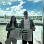 Opis szczegółowy zgodny z wymaganiami dostępności WCAG 2.2: Scena rozgrywa się w dzień, przy naturalnym świetle. Za dużą szybą w tle widać samoloty i terminal lotniczy. Niebo jest częściowo zachmurzone – białe, puszyste chmury kontrastują z błękitem nieba. Na pasie lotniska widoczny jest samolot z napisem „Copa Airlines” na kadłubie, ustawiony równolegle do okna. Na pierwszym planie: Stoi dwoje młodych ludzi, kobieta po lewej stronie i mężczyzna po prawej. Oboje mają na sobie casualowe, podróżne ubrania: jasne bluzy z kapturem i białe koszulki. Kobieta ma długie, falujące włosy w odcieniu brązu i nosi okulary. Ubrana jest w jasnoszare spodnie dresowe. Mężczyzna ma krótkie włosy i na głowie ciemną czapkę z daszkiem. Ma na sobie ciemne spodenki. Oboje trzymają wspólnie biało-szarą flagę z nadrukiem logo oraz napisem: „Fundacja Edukacji Rozwoju i Innowacji Staszica”. Logo składa się z liter F, E, R, I stylizowanych na geometryczne kształty w kolorze zielonym. Tło i otoczenie: Za nimi, przez wielką szybę, widać płyty postojowe lotniska oznaczone żółtymi liniami, zaparkowane samoloty, a dalej terminale i hangary. Na niebie przeważa jasne światło, co sprawia, że postacie są lekko przyciemnione w kontraście do tła. Podsumowanie dla osoby niewidomej: Zdjęcie przedstawia młodą kobietę i młodego mężczyznę stojących w terminalu lotniska przed dużym oknem, za którym widać samoloty i terminal. Oboje uśmiechają się lekko i trzymają flagę z napisem „Fundacja Edukacji Rozwoju i Innowacji Staszica”. Ich ubiór jest sportowy i wygodny, jak na podróż samolotem. Scena ma charakter spokojny, podróżny, z jasnym i pogodnym tłem.