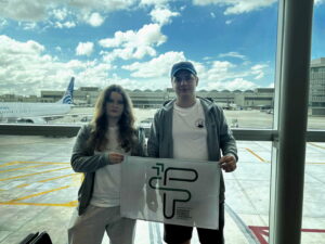 Opis szczegółowy zgodny z wymaganiami dostępności WCAG 2.2: Scena rozgrywa się w dzień, przy naturalnym świetle. Za dużą szybą w tle widać samoloty i terminal lotniczy. Niebo jest częściowo zachmurzone – białe, puszyste chmury kontrastują z błękitem nieba. Na pasie lotniska widoczny jest samolot z napisem „Copa Airlines” na kadłubie, ustawiony równolegle do okna. Na pierwszym planie: Stoi dwoje młodych ludzi, kobieta po lewej stronie i mężczyzna po prawej. Oboje mają na sobie casualowe, podróżne ubrania: jasne bluzy z kapturem i białe koszulki. Kobieta ma długie, falujące włosy w odcieniu brązu i nosi okulary. Ubrana jest w jasnoszare spodnie dresowe. Mężczyzna ma krótkie włosy i na głowie ciemną czapkę z daszkiem. Ma na sobie ciemne spodenki. Oboje trzymają wspólnie biało-szarą flagę z nadrukiem logo oraz napisem: „Fundacja Edukacji Rozwoju i Innowacji Staszica”. Logo składa się z liter F, E, R, I stylizowanych na geometryczne kształty w kolorze zielonym. Tło i otoczenie: Za nimi, przez wielką szybę, widać płyty postojowe lotniska oznaczone żółtymi liniami, zaparkowane samoloty, a dalej terminale i hangary. Na niebie przeważa jasne światło, co sprawia, że postacie są lekko przyciemnione w kontraście do tła. Podsumowanie dla osoby niewidomej: Zdjęcie przedstawia młodą kobietę i młodego mężczyznę stojących w terminalu lotniska przed dużym oknem, za którym widać samoloty i terminal. Oboje uśmiechają się lekko i trzymają flagę z napisem „Fundacja Edukacji Rozwoju i Innowacji Staszica”. Ich ubiór jest sportowy i wygodny, jak na podróż samolotem. Scena ma charakter spokojny, podróżny, z jasnym i pogodnym tłem.