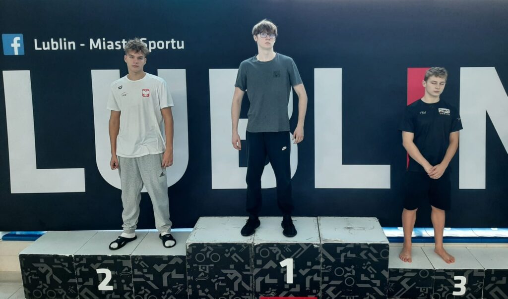 zdjęcie przedstawia trzech młodych mężczyzn stojących na podium w tym samym miejscu — w tle również znajduje się czarna tablica z napisem „Lublin – Miasto Sportu”. Na najwyższym stopniu podium, oznaczonym cyfrą 1, stoi chłopak w ciemnym szarym T-shircie i czarnych dresach. Ma krótkie włosy, nosi okulary i stoi prosto, z rękami po bokach. Po jego lewej stronie (miejsce drugie, z cyfrą 2) stoi chłopak w białej koszulce sportowej z godłem Polski i szarych spodniach dresowych. Na nogach ma czarne klapki z białymi paskami. Po prawej stronie (miejsce trzecie, z cyfrą 3) stoi chłopak w czarnej koszulce sportowej z napisem „Lublinianka” lub „Lublin” (napis częściowo widoczny) i czarnych spodenkach. Stoi boso, z rękami splecionymi przed sobą i głową lekko pochyloną w dół. Podium jest czarne z białymi cyframi i geometrycznymi wzorami. Scena przedstawia zakończenie zawodów sportowych, najprawdopodobniej pływackich. Chłopcy stoją spokojnie, pozując do zdjęcia pamiątkowego po ogłoszeniu wyników. Tło i oświetlenie są takie same jak na pierwszym zdjęciu – jasne, z refleksami światła odbijającego się od przeszklonych ścian basenu.
