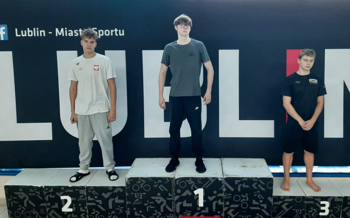 zdjęcie przedstawia trzech młodych mężczyzn stojących na podium w tym samym miejscu — w tle również znajduje się czarna tablica z napisem „Lublin – Miasto Sportu”. Na najwyższym stopniu podium, oznaczonym cyfrą 1, stoi chłopak w ciemnym szarym T-shircie i czarnych dresach. Ma krótkie włosy, nosi okulary i stoi prosto, z rękami po bokach. Po jego lewej stronie (miejsce drugie, z cyfrą 2) stoi chłopak w białej koszulce sportowej z godłem Polski i szarych spodniach dresowych. Na nogach ma czarne klapki z białymi paskami. Po prawej stronie (miejsce trzecie, z cyfrą 3) stoi chłopak w czarnej koszulce sportowej z napisem „Lublinianka” lub „Lublin” (napis częściowo widoczny) i czarnych spodenkach. Stoi boso, z rękami splecionymi przed sobą i głową lekko pochyloną w dół. Podium jest czarne z białymi cyframi i geometrycznymi wzorami. Scena przedstawia zakończenie zawodów sportowych, najprawdopodobniej pływackich. Chłopcy stoją spokojnie, pozując do zdjęcia pamiątkowego po ogłoszeniu wyników. Tło i oświetlenie są takie same jak na pierwszym zdjęciu – jasne, z refleksami światła odbijającego się od przeszklonych ścian basenu.