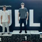 zdjęcie przedstawia trzech młodych mężczyzn stojących na podium w tym samym miejscu — w tle również znajduje się czarna tablica z napisem „Lublin – Miasto Sportu”. Na najwyższym stopniu podium, oznaczonym cyfrą 1, stoi chłopak w ciemnym szarym T-shircie i czarnych dresach. Ma krótkie włosy, nosi okulary i stoi prosto, z rękami po bokach. Po jego lewej stronie (miejsce drugie, z cyfrą 2) stoi chłopak w białej koszulce sportowej z godłem Polski i szarych spodniach dresowych. Na nogach ma czarne klapki z białymi paskami. Po prawej stronie (miejsce trzecie, z cyfrą 3) stoi chłopak w czarnej koszulce sportowej z napisem „Lublinianka” lub „Lublin” (napis częściowo widoczny) i czarnych spodenkach. Stoi boso, z rękami splecionymi przed sobą i głową lekko pochyloną w dół. Podium jest czarne z białymi cyframi i geometrycznymi wzorami. Scena przedstawia zakończenie zawodów sportowych, najprawdopodobniej pływackich. Chłopcy stoją spokojnie, pozując do zdjęcia pamiątkowego po ogłoszeniu wyników. Tło i oświetlenie są takie same jak na pierwszym zdjęciu – jasne, z refleksami światła odbijającego się od przeszklonych ścian basenu.