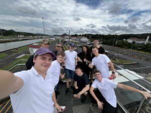 Grupa dziesięciu młodych osób stoi na metalowej platformie z widokiem na śluzy kanału wodnego. W tle widać system śluz, tory, budynki techniczne i zieleń wzgórz. Osoby uśmiechają się do aparatu, część pokazuje kciuki w górę. Zdjęcie wykonane z góry w słoneczny, lekko pochmurny dzień. Kolory: dominują biele koszulek, stalowy metal platformy i zieleń krajobrazu.